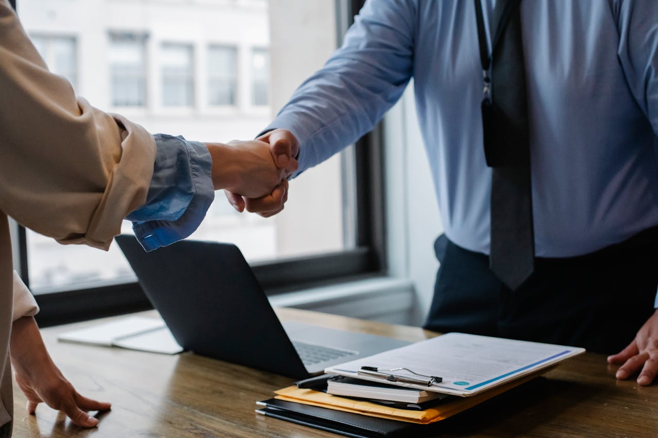 Two people in business attire shake hands over a desk with a laptop, clipboard, and paperwork, suggesting a professional or business agreement in an office setting.