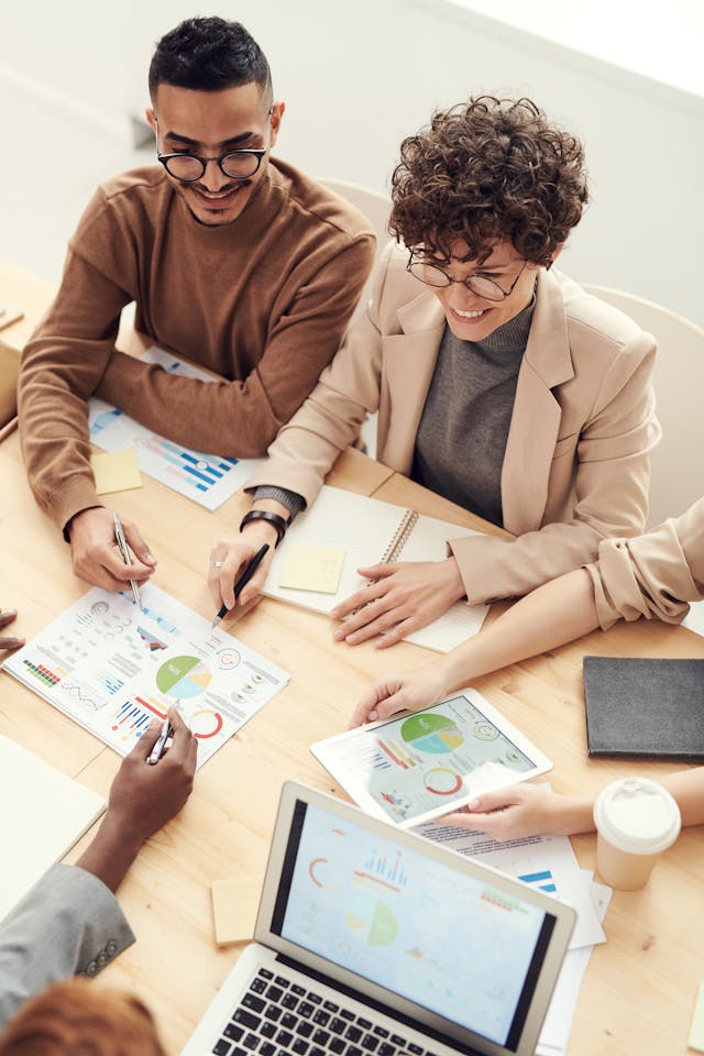 Four people sit at a wooden table, discussing colorful charts and graphs. A laptop, notebooks, documents, and a coffee cup are on the table, suggesting a collaborative business meeting or brainstorming session.