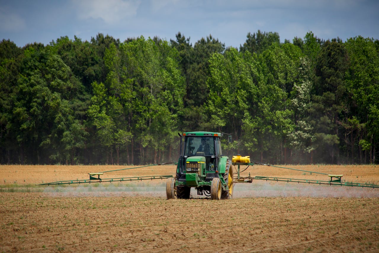 A green tractor sprays crops in a large, open field with sparse vegetation, bordered by dense green trees under a partly cloudy sky.