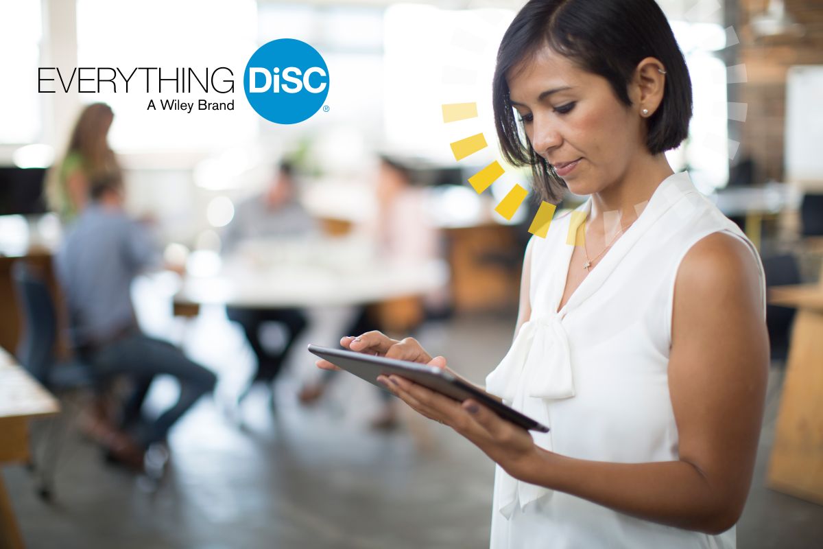 A woman in a white sleeveless blouse uses a tablet in a modern office. The "Everything DiSC" logo is in the upper left corner. Several people are working in the blurred background.