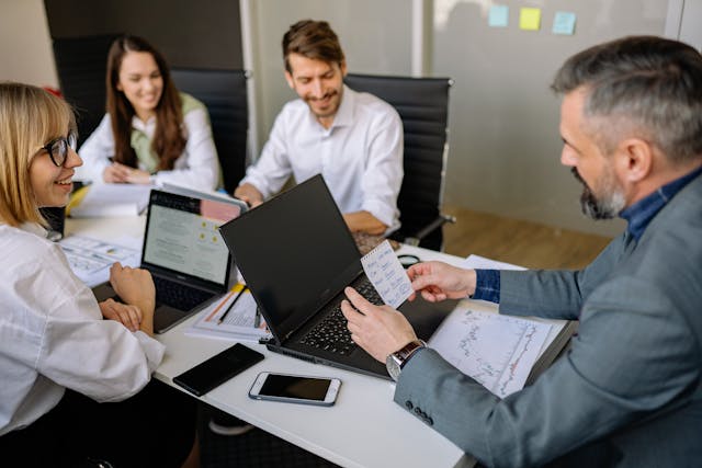 Four people sit around a table in an office, discussing documents and laptops. One man shows a paper to the group while others smile and engage in conversation. A smartphone and notebooks are on the table.