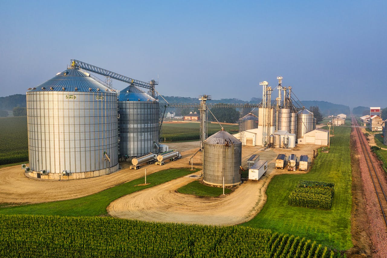 Aerial view of a grain storage facility with large metal silos, trucks, and farm buildings, surrounded by green fields and bordered by a railroad track under a clear sky.
