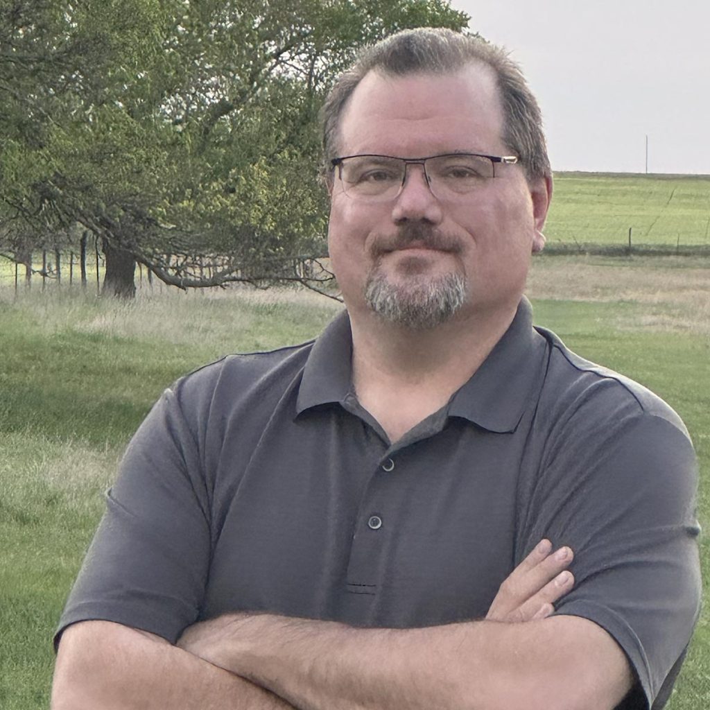 Nick crosses his arms in a grey polo, standing in front of a green field with a wide tree in the background. Square orientation