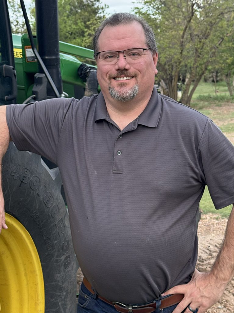 Nick leans his right elbow on the wheel of a tractor while the other hands rests on his hip. He's wearing a grey polo, and the picture has a tall portrait orientation