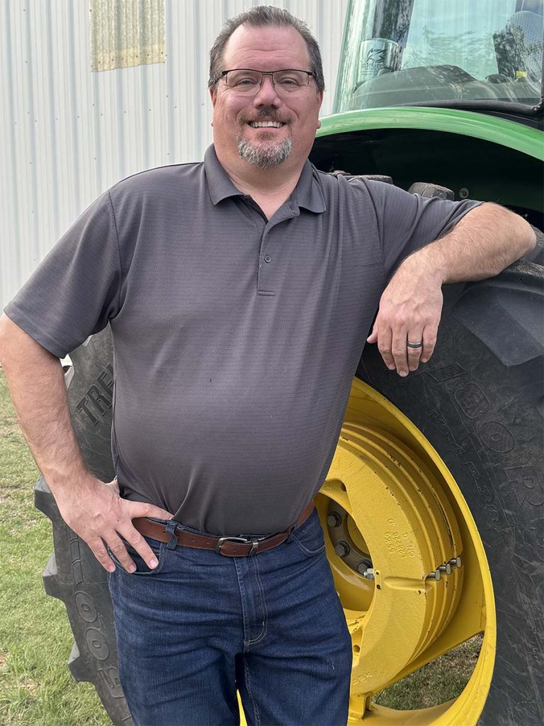 Nick leans his left elbow on the wheel of a tractor while the other hands rests on his hip. He's wearing a grey polo, and the picture has a tall portrait orientation