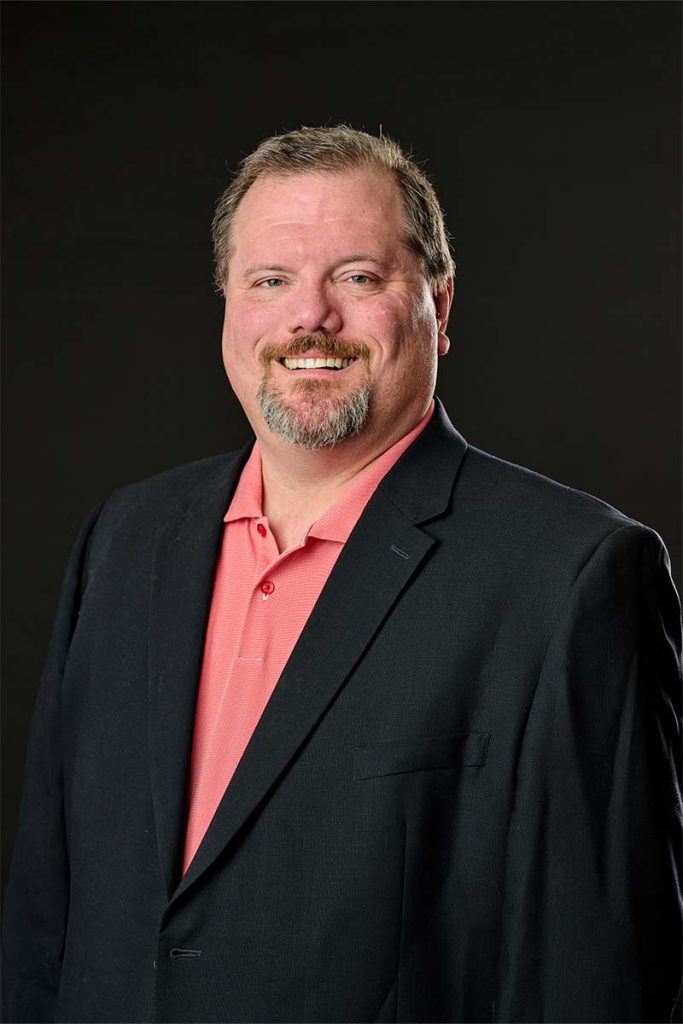 Nick smiling in a salmon dress shirt and charcoal suite jacket on a black background. Tall portrait orientation