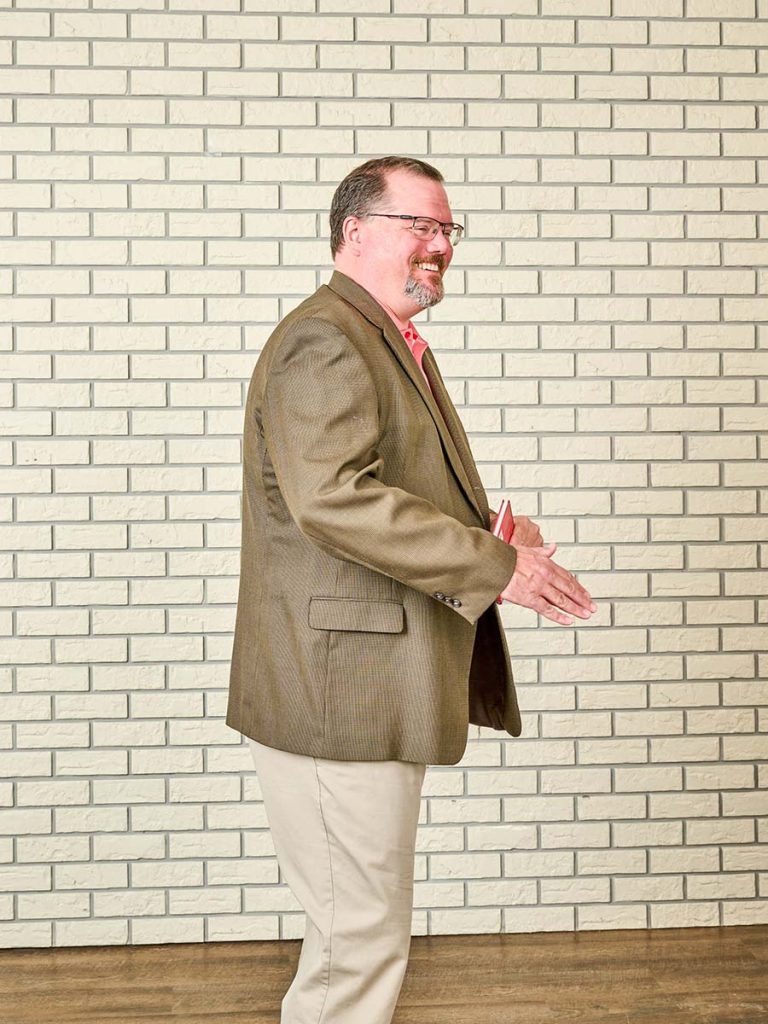 Nick standing in front of a beige brick background in a dark beige jacket, khaki pants, and a salmon dress shirt