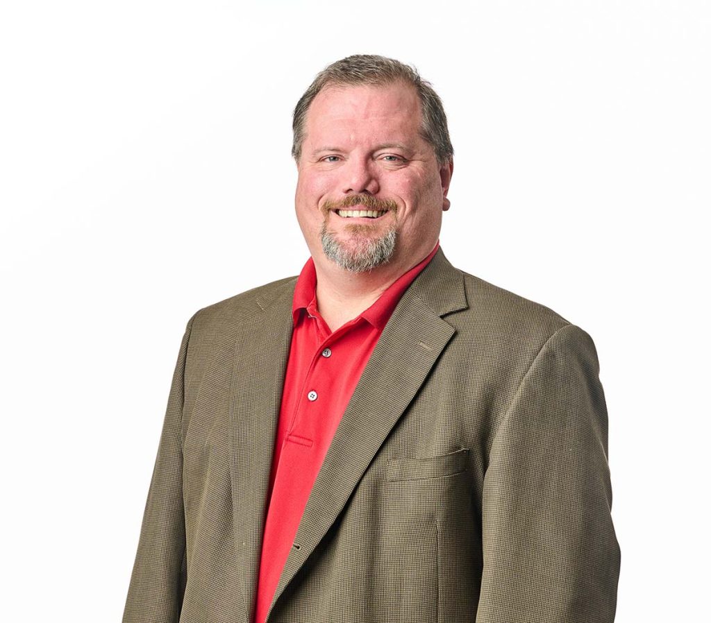 Nick smiling in a red polo and beige suite jacket on a white background. Relatively square orientation
