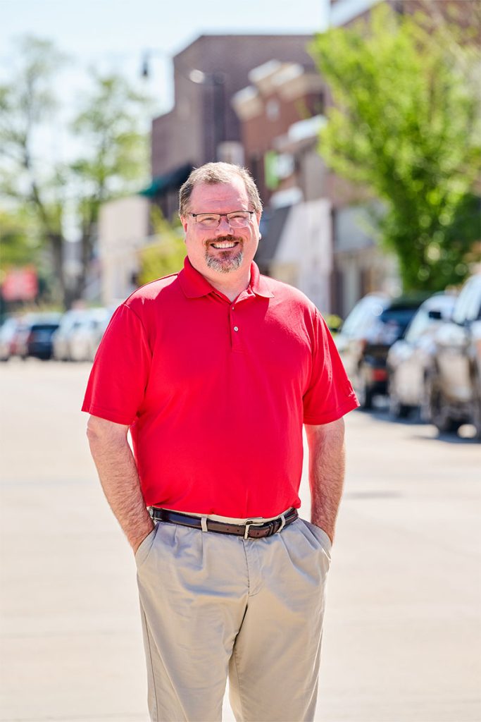 Nick smiling with his hands in his pockets, wearing a red polo in front of a shopping street background. Tall portrait orientation