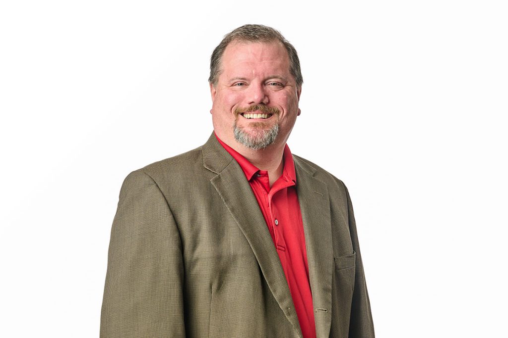 Nick smiling in a red polo and beige suite jacket on a white background. Landscape orientation