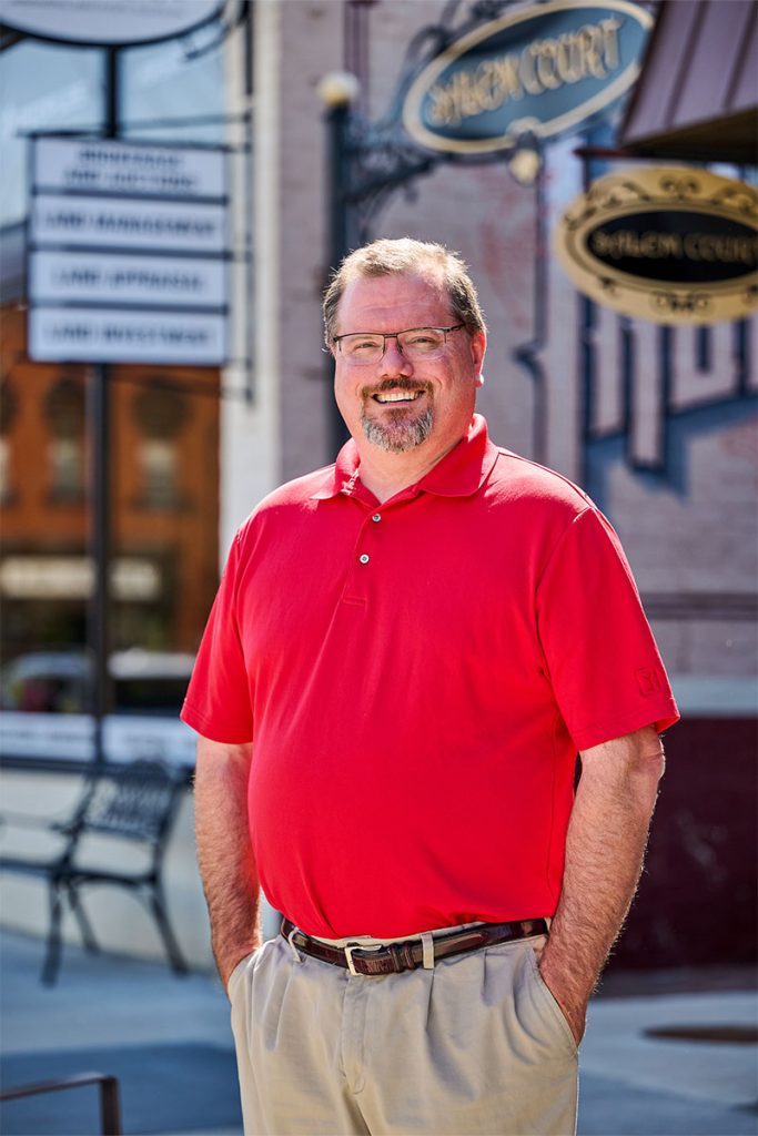 Nick smiling with his hands in his pockets, wearing a red polo in front of a brick facade shop background. Tall portrait orientation