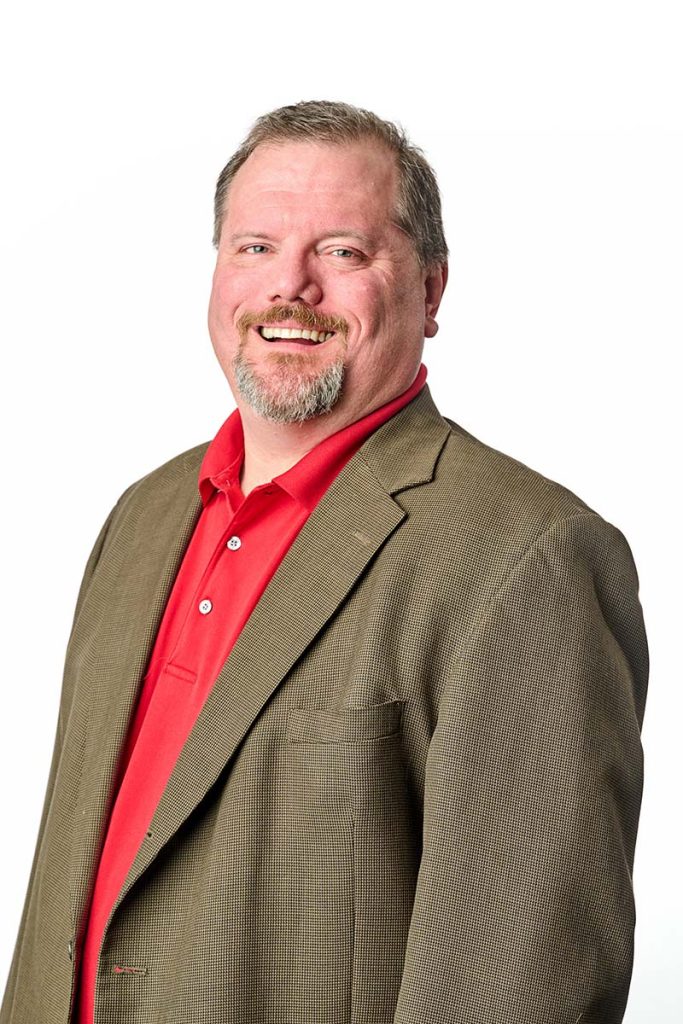 Nick smiling in a red polo and beige suite jacket on a white background. Tall portrait orientation.